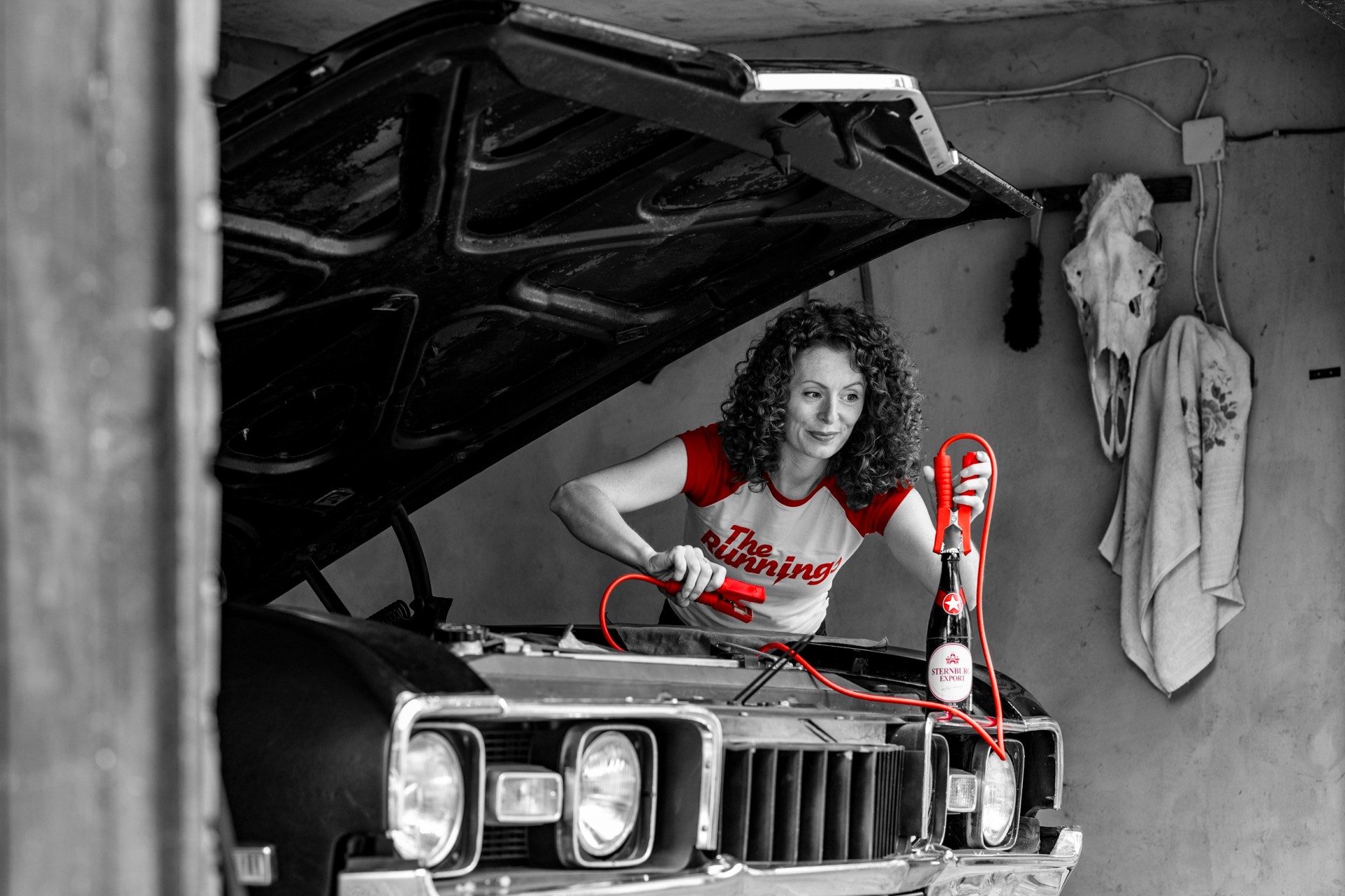 Frau hält in einer Garage an einem offenen Motorraum ein Überbrückungskabel an eine Sternburg Bierflasche. An der Wand hängen Tierschädel und Handtuch.