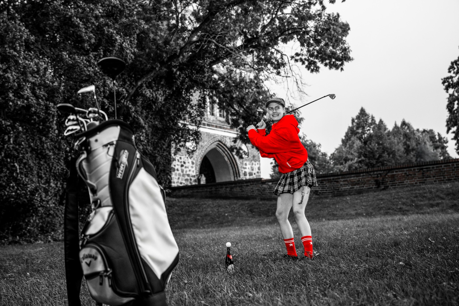 Person mit Golfschläger, die einen roten Pullover und rote Strümpfe trägt, auf einer Wiese neben einer Sternburg-Bierflasche.