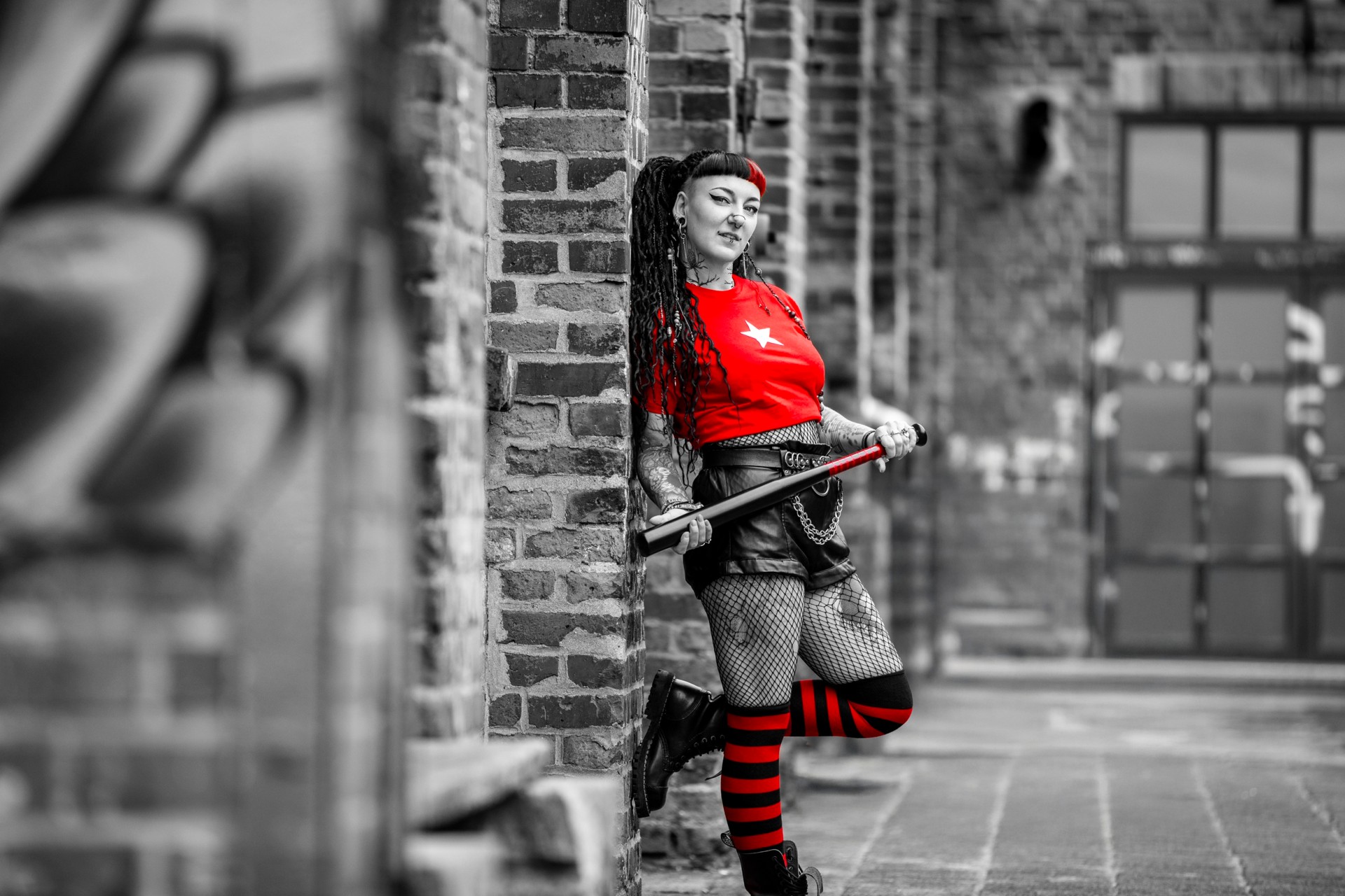 Frau mit rotem Shirt lehnt an Backsteinwand und hält Baseballschläger. Im Hintergrund Industriehof mit Tor und Pflaster.