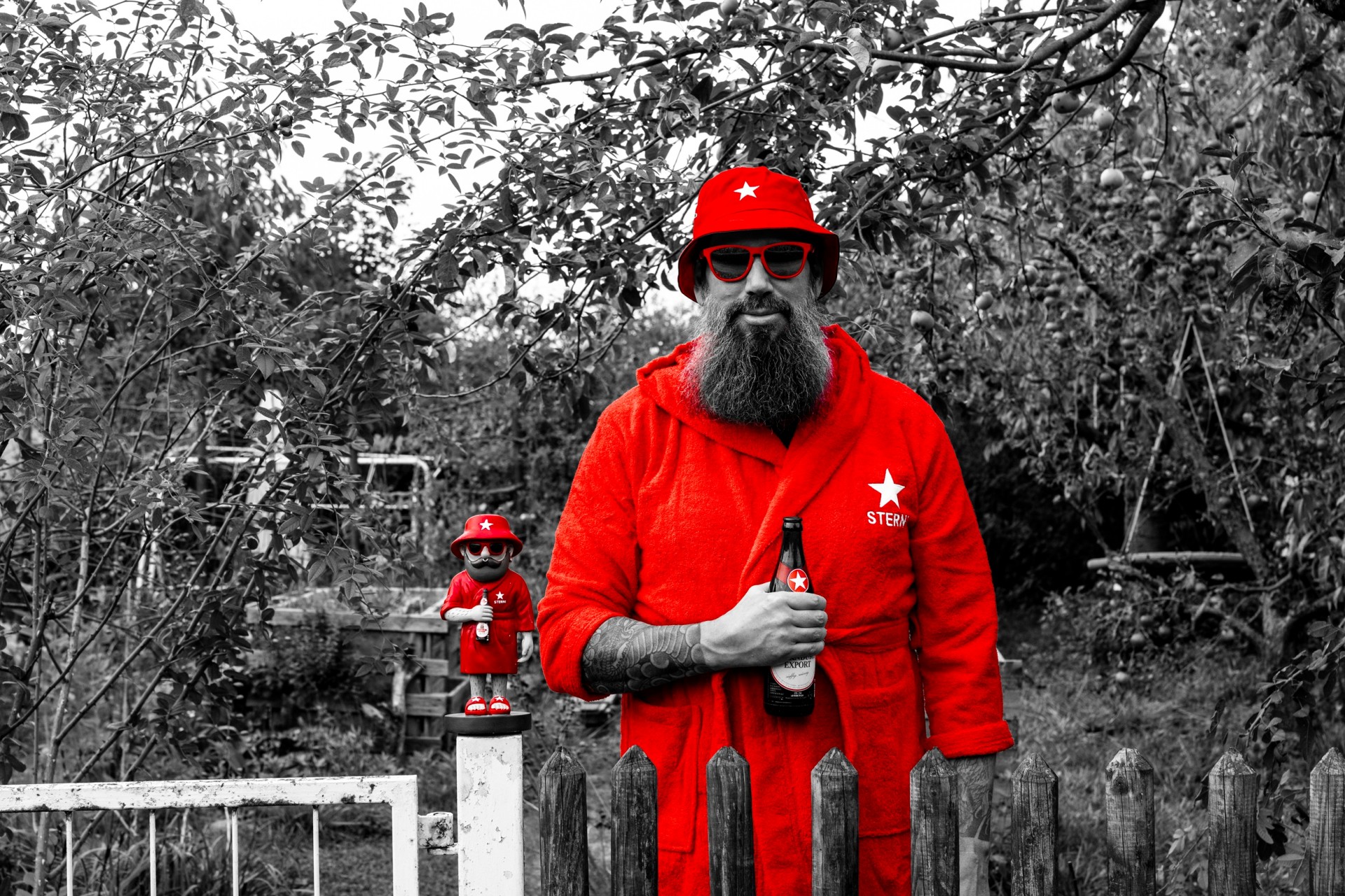 Mann in rotem Bademantel und Hut hält Sterni-Bierflasche, steht neben passender Figur in Garten mit Zaun.