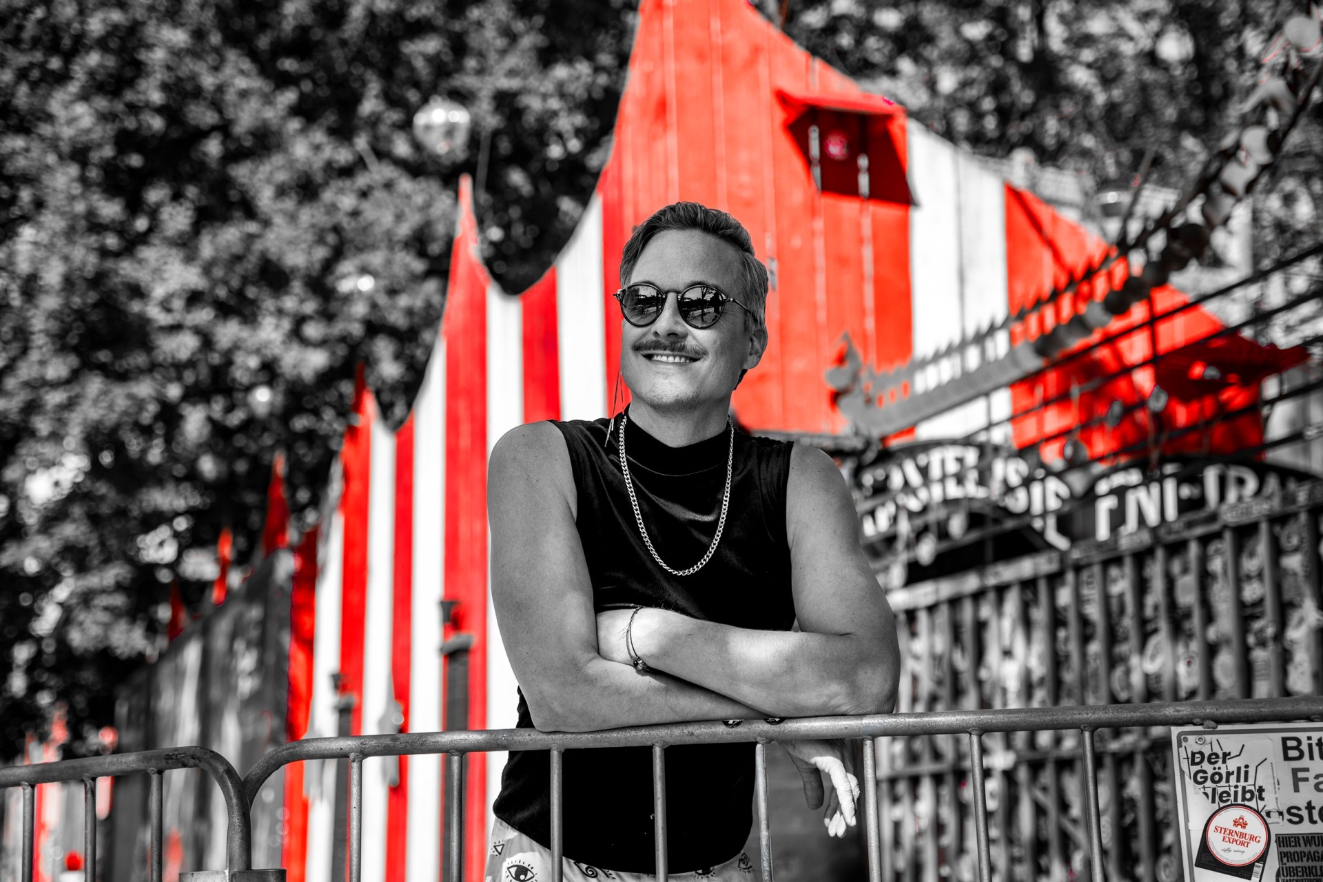 Mann mit Sonnenbrille lehnt mit verschränkten Armen an einem Gitter. Rot und weiß gestreiftes Gebäude im Hintergrund.
