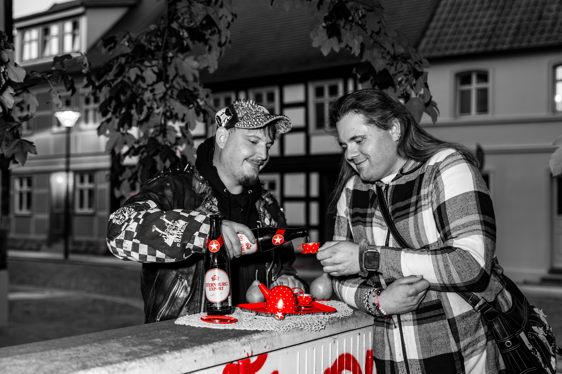 Zwei Männer stehen abends lachend in einer Altstadt. Sie trinken aus einem roten Teeservice und füllen sich Bier aus einer Sternburg-Flasche in die kleine Teetasse.
