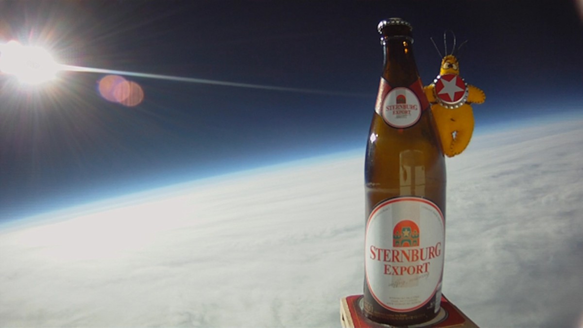 Eine Bierflasche steht im Vordergrund mit einer Comicfigur daneben vor der Erdkrümmung im All.