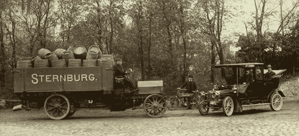 Zwei alte Fahrzeuge stehen auf einer Allee, eines ist ein Lastwagen voller Fässer, das andere ein Auto.