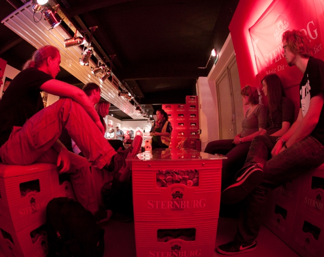 Mehrere Personen sitzen entspannt auf roten Kisten in einem beleuchteten Raum.