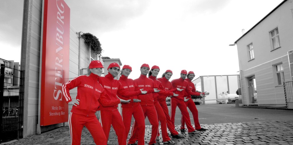 Sieben Personen in roten Overalls und Mützen stehen auf einem gepflasterten Hof.