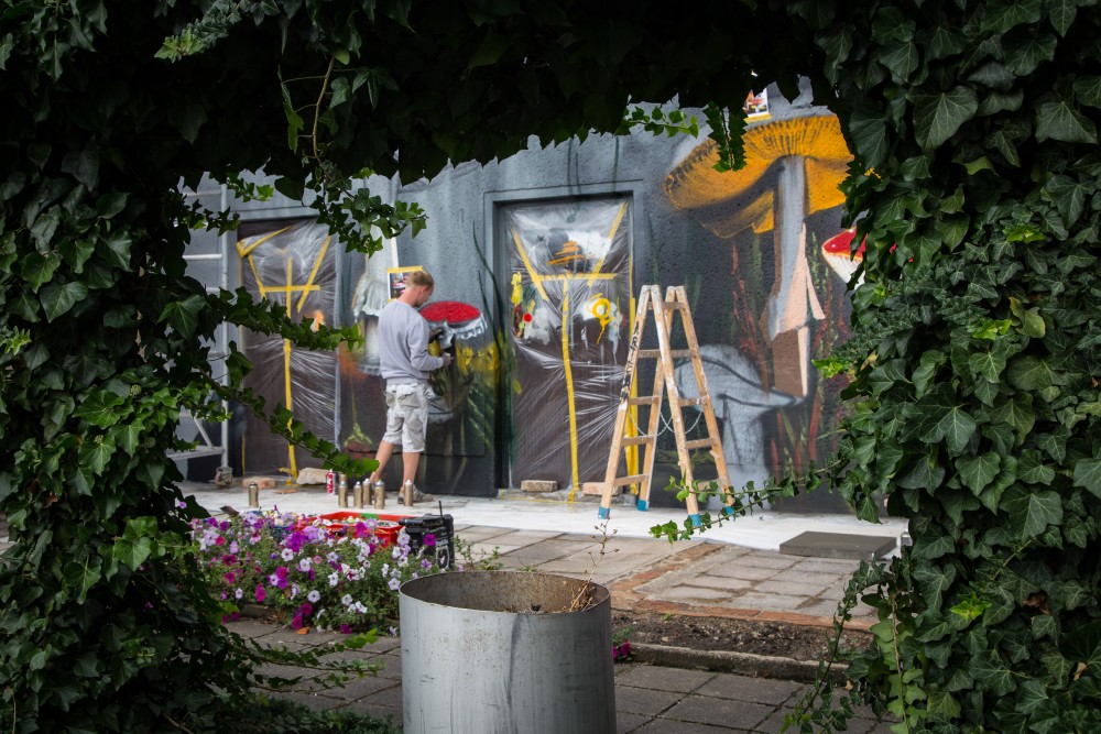 Ein Künstler bemalt ein großes Graffiti-Wandbild mit Pilzen auf eine graue Mauer, während grüner Weinranken im Vordergrund das Bild umrahmen.