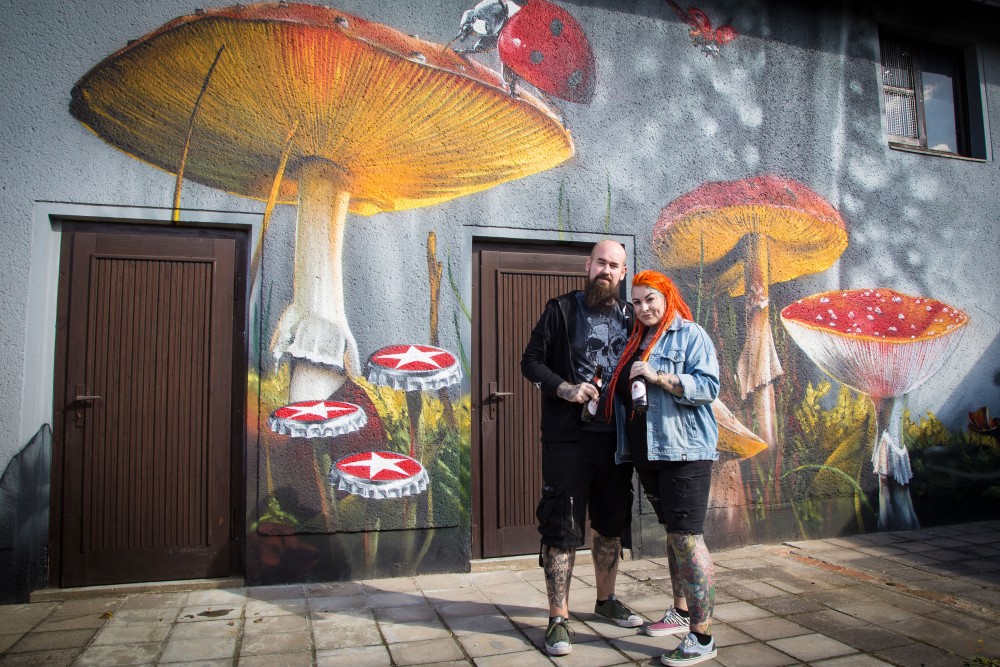 Malerische Graffiti-Wandgestaltung mit riesigen Pilzen neben zwei Personen vor einem grauen Gebäude. Ein Mann und eine Frau posieren.