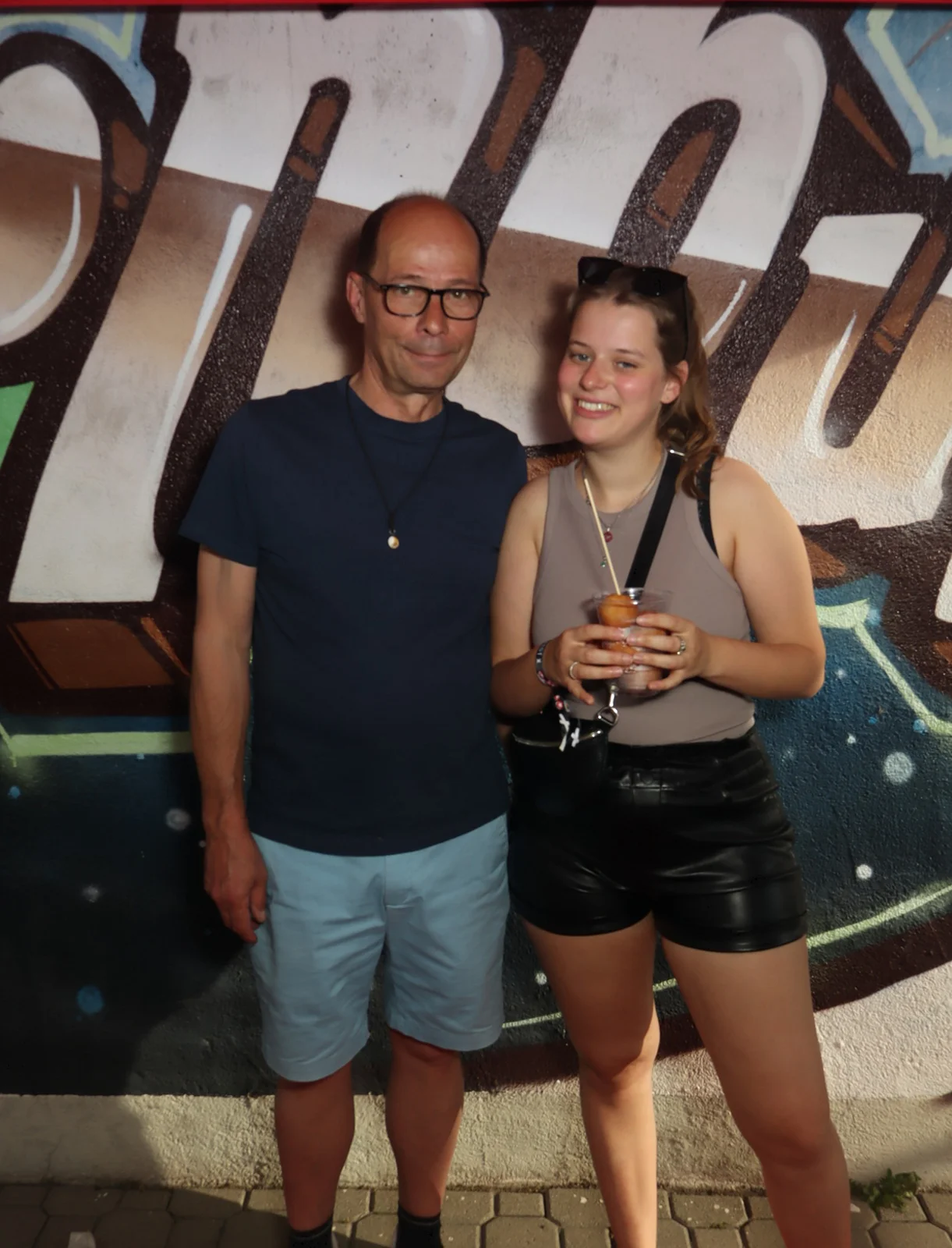  Ein Mann und eine junge Frau stehen nebeneinander. Die Frau hält ein Getränk, im Hintergrund eine bemalte Wand.