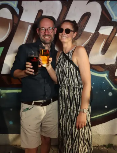 Eine Frau im weißen Kleid mit Muster und ein Mann mit Hut stehen nebeneinander. Im Hintergrund eine Wand mit Graffiti.