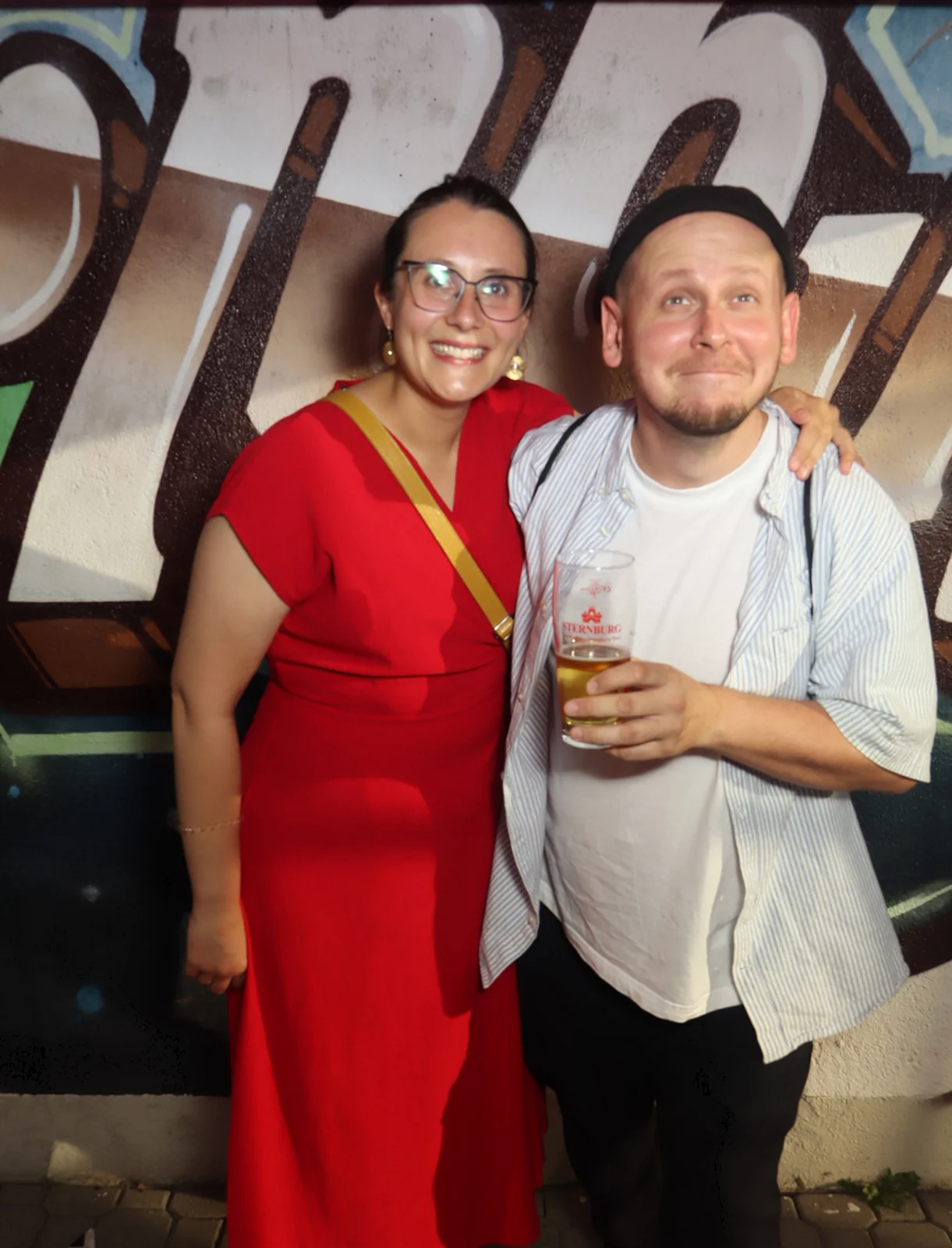 Ein Mann und eine Frau stehen eng beieinander und lächeln. Im Hintergrund eine mit Buchstaben bemalte Wand.