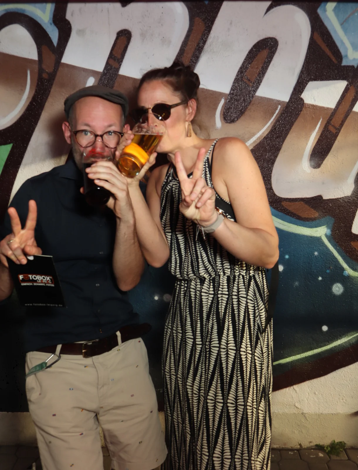  Eine Frau in geblümtem Kleid und ein Mann mit Bart stehen nebeneinander. Im Hintergrund eine Graffitiwand.