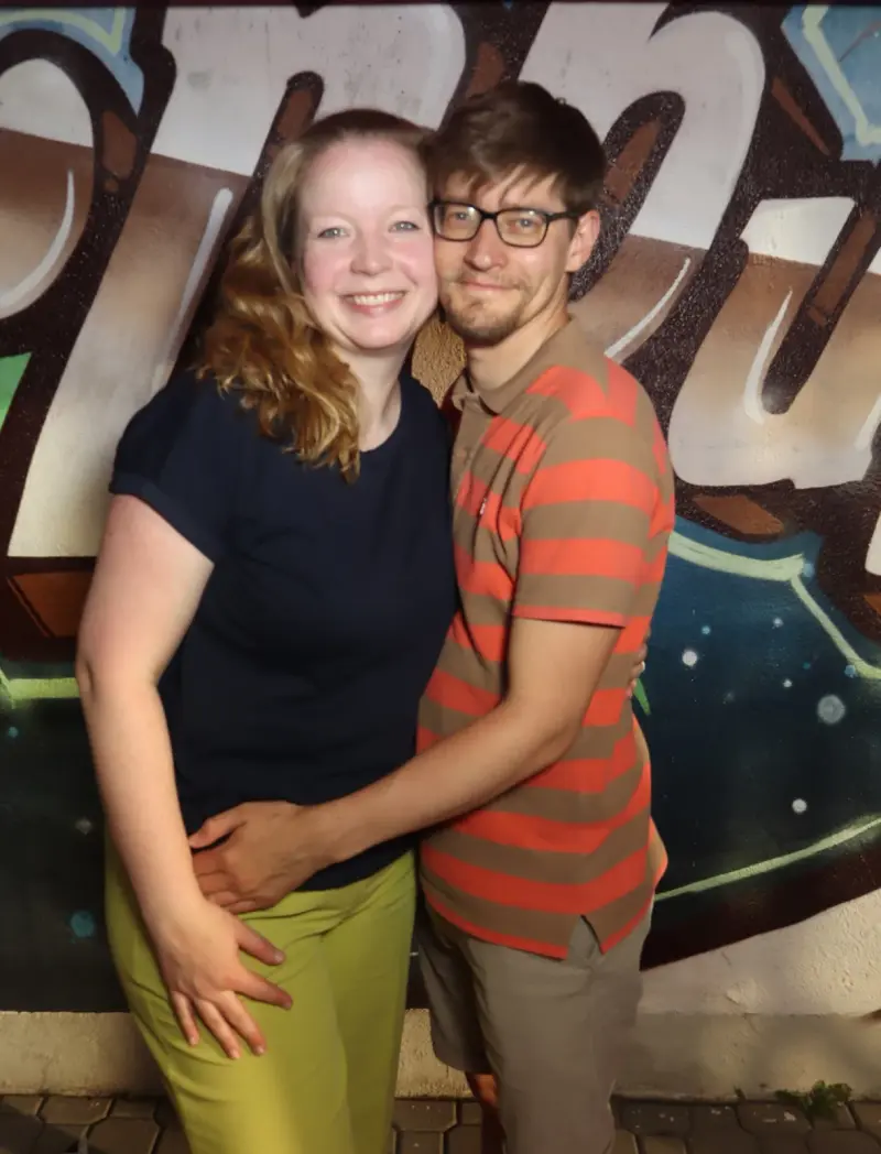 Ein Paar steht eng umarmt vor einer bemalten Wand mit großflächigem Graffiti-Schriftzug.