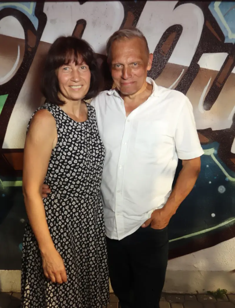 Ein Paar steht eng beieinander, die Frau trägt ein geblümtes Kleid, der Mann ein weißes Hemd. Der Hintergrund zeigt Graffiti.