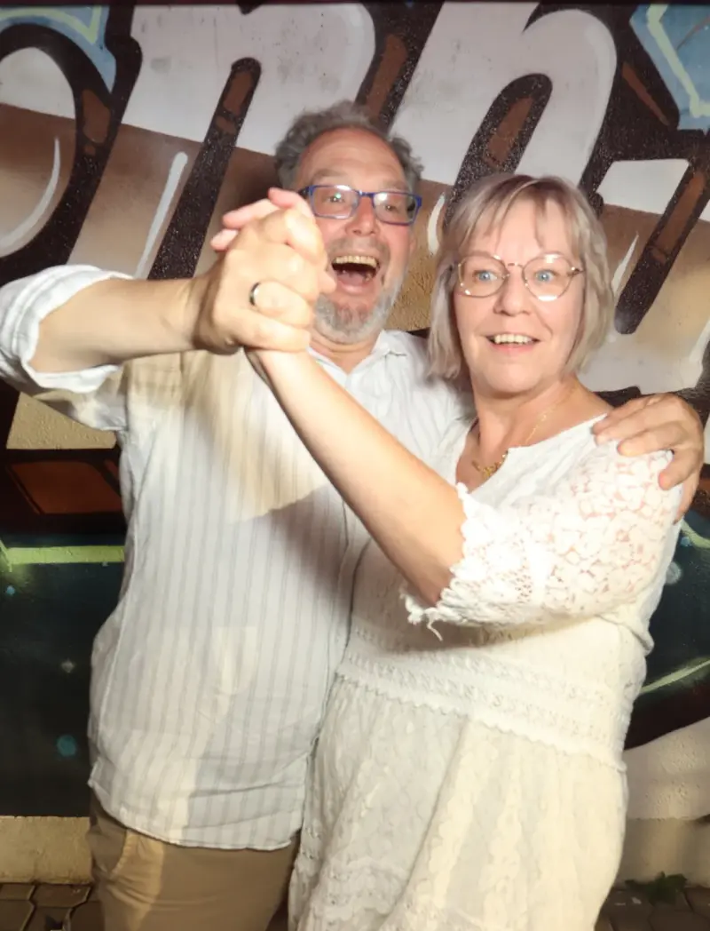 Ein älteres Paar tanzt eng umschlungen, beide tragen helle Kleidung. Im Hintergrund ist eine Graffiti Wand zu sehen.