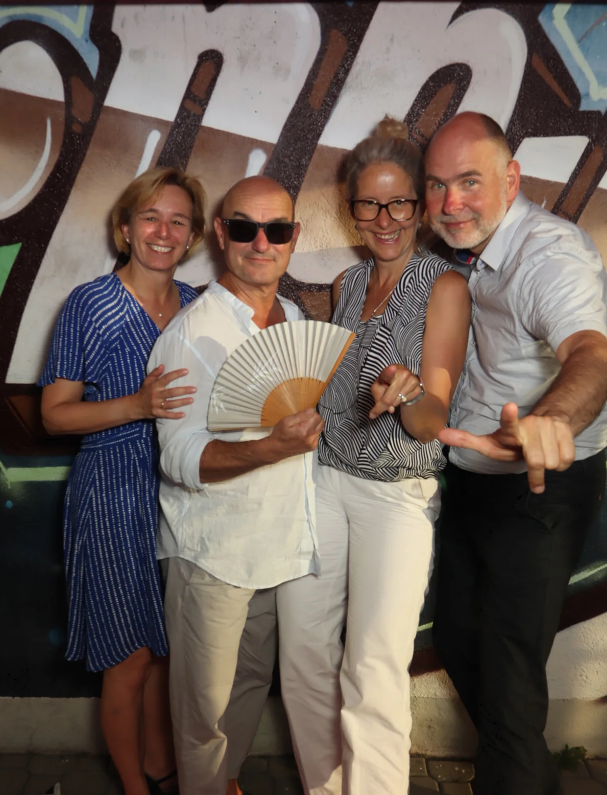Vier Erwachsene posieren nah beieinander, eine Person hält einen Fächer. Im Hintergrund befindet sich eine Graffiti-Wand.