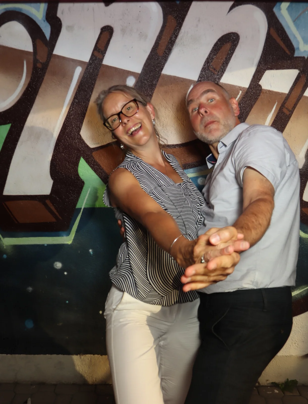 Ein Paar tanzt eng umschlungen, die Frau trägt eine Brille und gestreiftes Oberteil. Der Hintergrund zeigt bunte Graffiti.