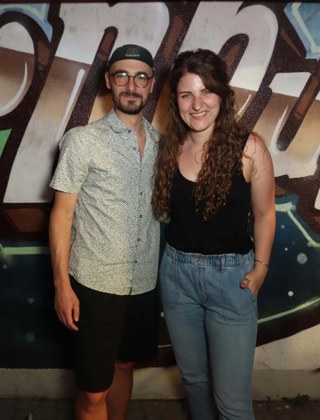 Ein junges Paar steht nebeneinander, der Mann trägt Brille, die Frau hat lange Locken. Der Hintergrund zeigt Graffiti.