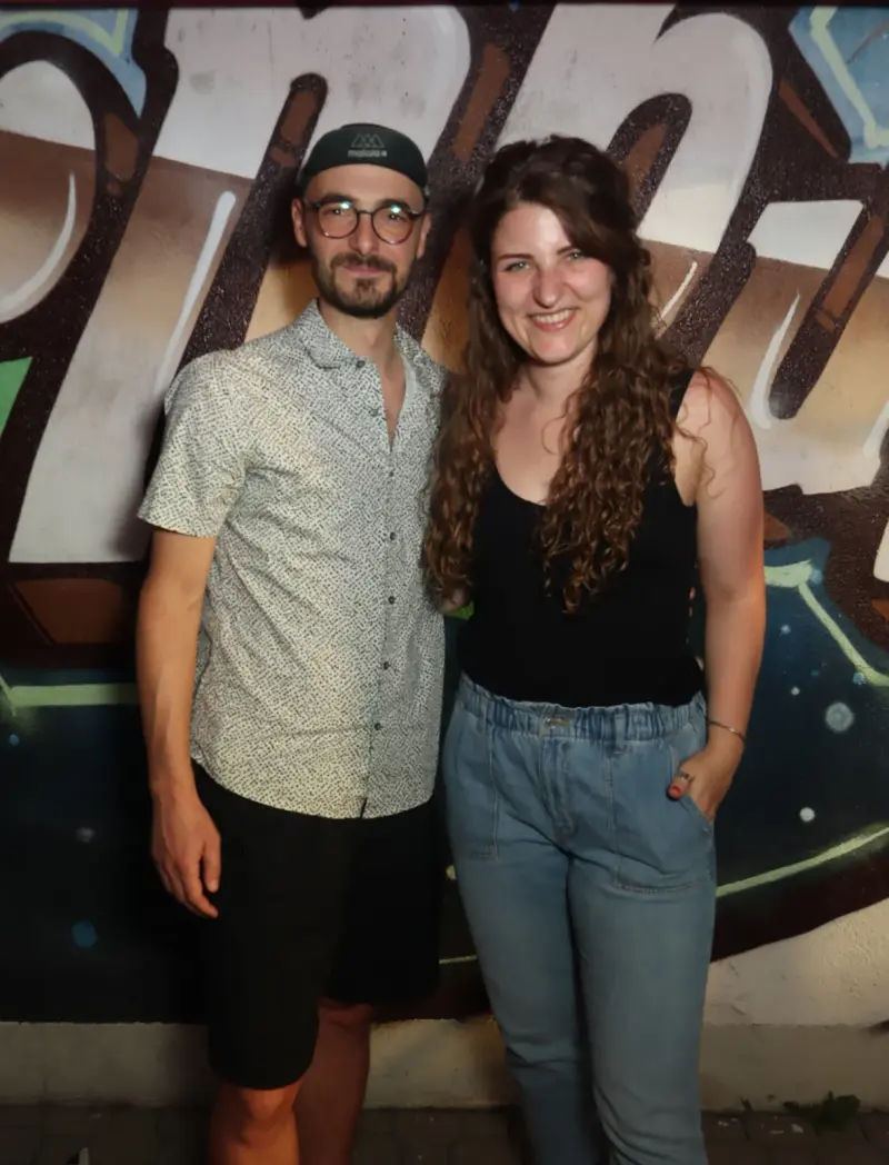 Ein junges Paar steht nebeneinander, der Mann trägt Brille, die Frau hat lange Locken. Der Hintergrund zeigt Graffiti.