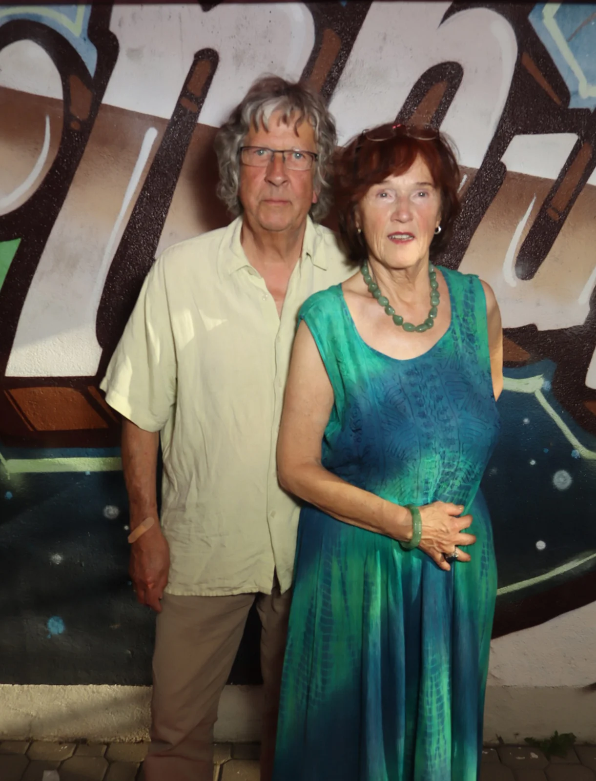 Ein älteres Paar steht vor einer bemalten Wand. Die Frau trägt ein langes blau-grünes Kleid und eine grüne Kette.