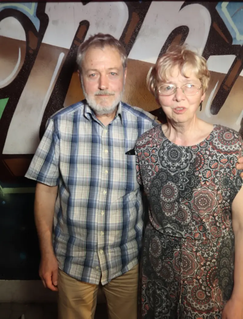 Ein älteres Paar steht nebeneinander, der Mann trägt kariertes Hemd, die Frau ein gemustertes Kleid. Im Hintergrund Graffiti.