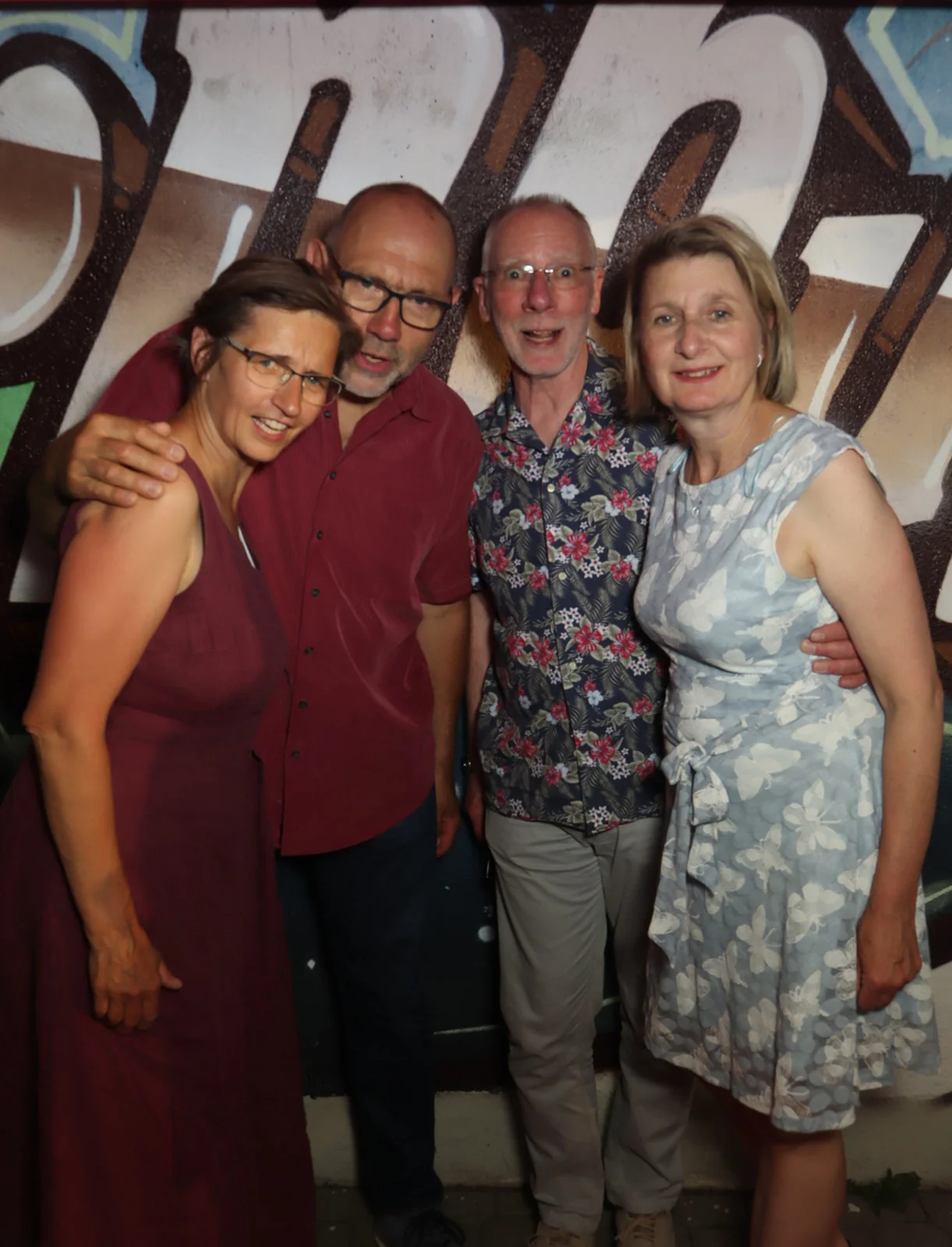 Vier Erwachsene stehen dicht beisammen, zwei tragen Brillen. Der Hintergrund zeigt eine bunt bemalte Wand mit Graffiti.