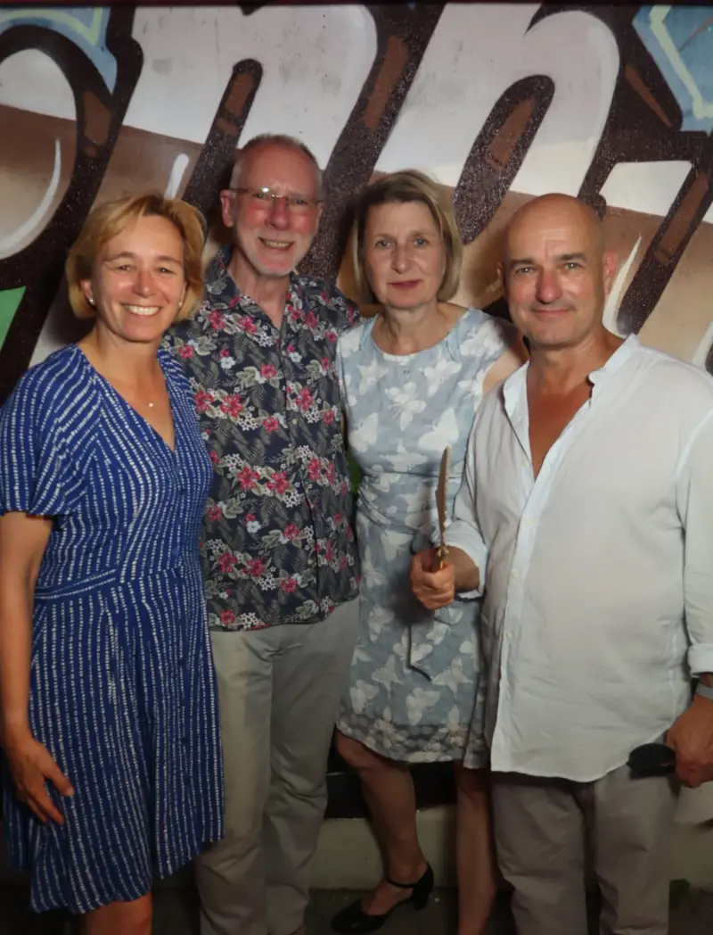 Vier Erwachsene stehen nebeneinander. Der Hintergrund zeigt eine bemalte Wand mit Graffiti.