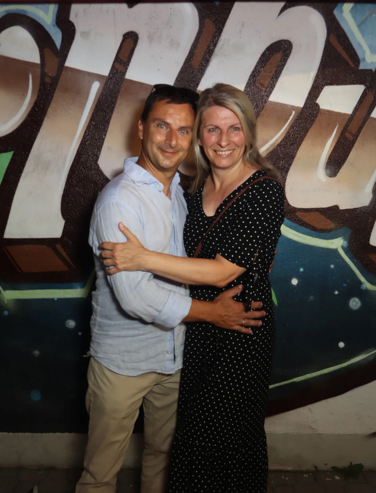 Ein Paar steht eng umschlungen, die Frau trägt ein gepunktetes Kleid. Der Hintergrund zeigt eine Wand mit buntem Graffiti.