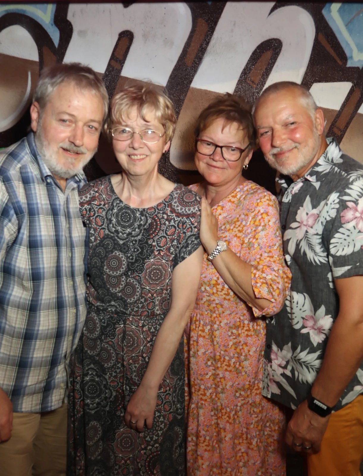 Zwei Paare stehen nebeneinander, alle Personen tragen gemusterte Kleidung. Der Hintergrund zeigt eine Wand mit großem Graffiti.