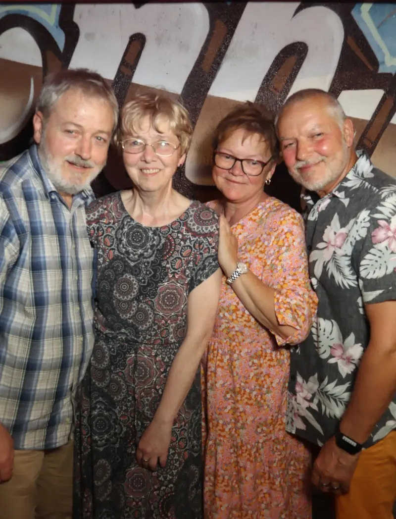 Zwei Paare stehen nebeneinander, alle Personen tragen gemusterte Kleidung. Der Hintergrund zeigt eine Wand mit großem Graffiti.