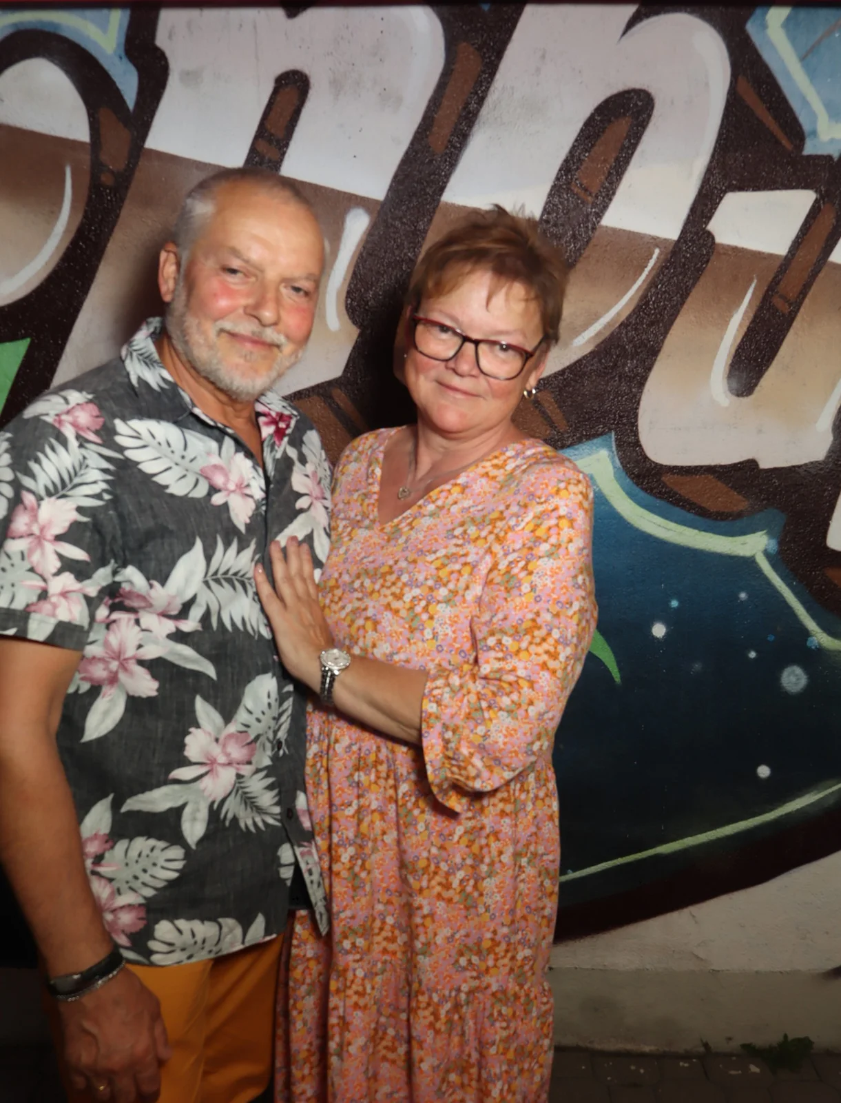 Ein Paar steht nebeneinander, der Mann trägt ein Hemd mit Blumenmuster, die Frau ein Kleid. Im Hintergrund Graffiti.