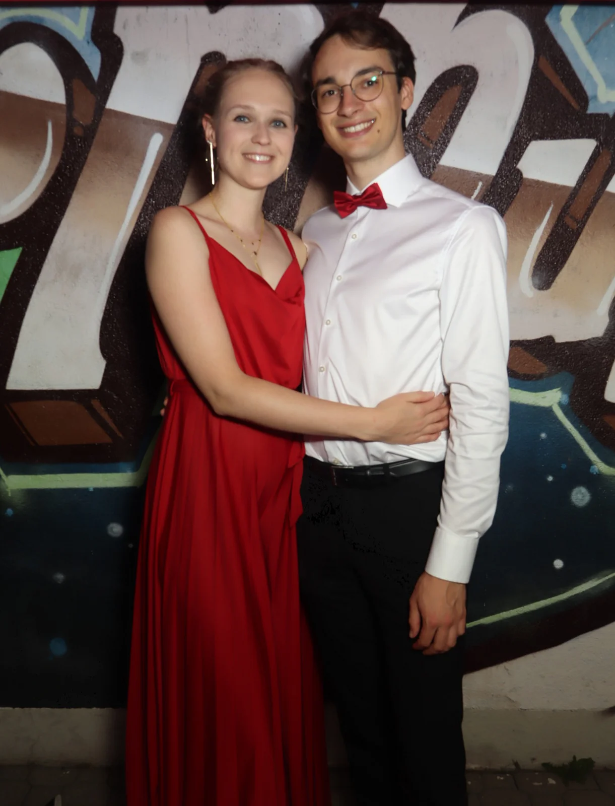 Ein junges Paar steht nebeneinander, die Frau trägt ein rotes Kleid, der Mann ein Hemd mit Fliege. Im Hintergrund Graffiti.
