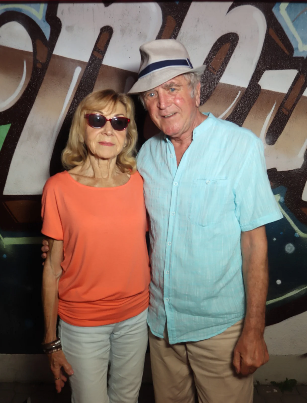 Ein älteres Paar steht nebeneinander, die Frau trägt Sonnenbrille, der Mann einen Hut. Im Hintergrund eine Wand mit Graffiti.