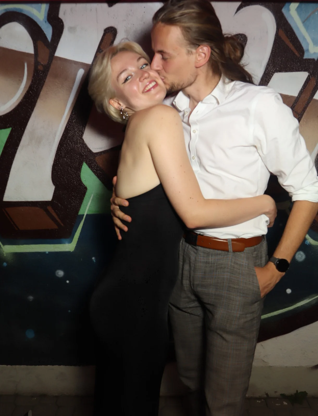 Ein junges Paar umarmt sich, der Mann küsst die Frau auf die Wange. Im Hintergrund eine bemalte Wand mit Graffiti.
