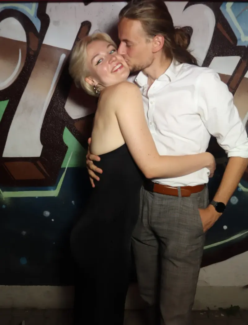 Ein junges Paar umarmt sich, der Mann küsst die Frau auf die Wange. Im Hintergrund eine bemalte Wand mit Graffiti.