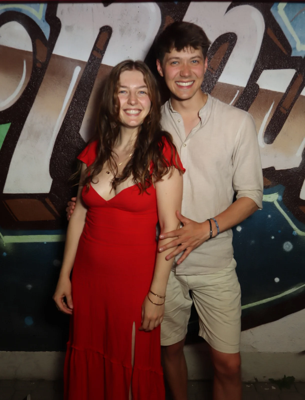 Ein junges Paar steht nebeneinander, die Frau trägt ein rotes Kleid mit Schlitz. Im Hintergrund eine Wand mit Graffiti.