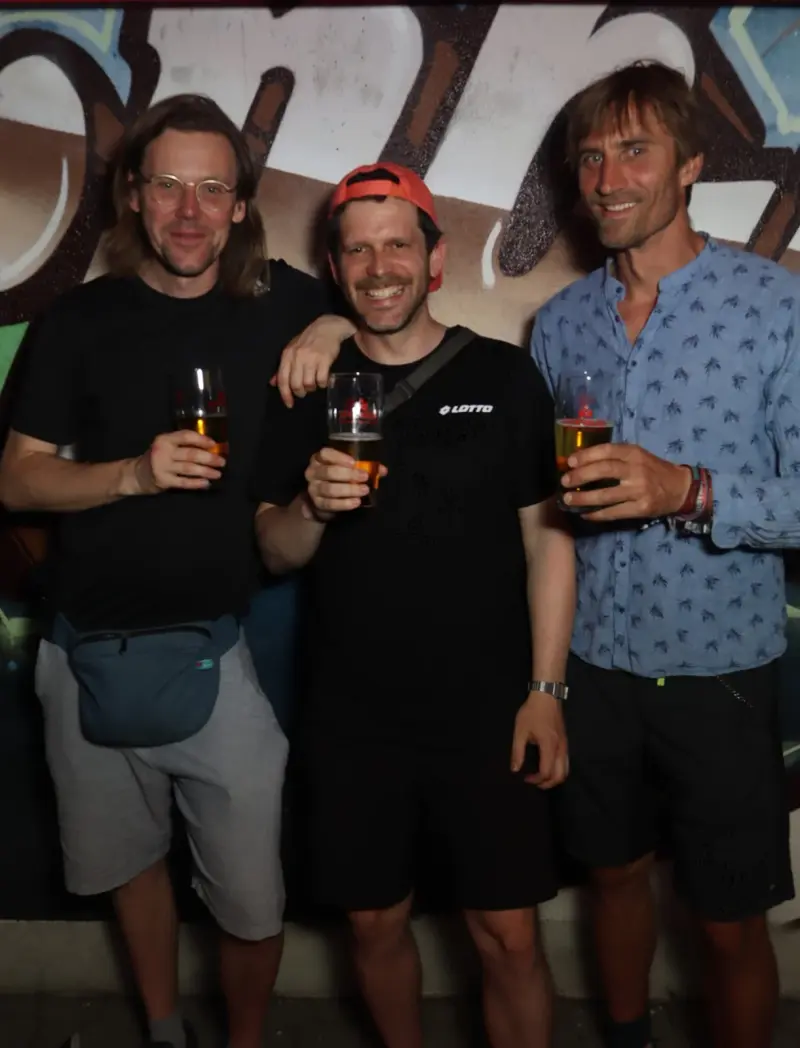 Drei Männer stehen nebeneinander und halten Biergläser in der Hand. Im Hintergrund ist eine bemalte Wand mit Graffiti.