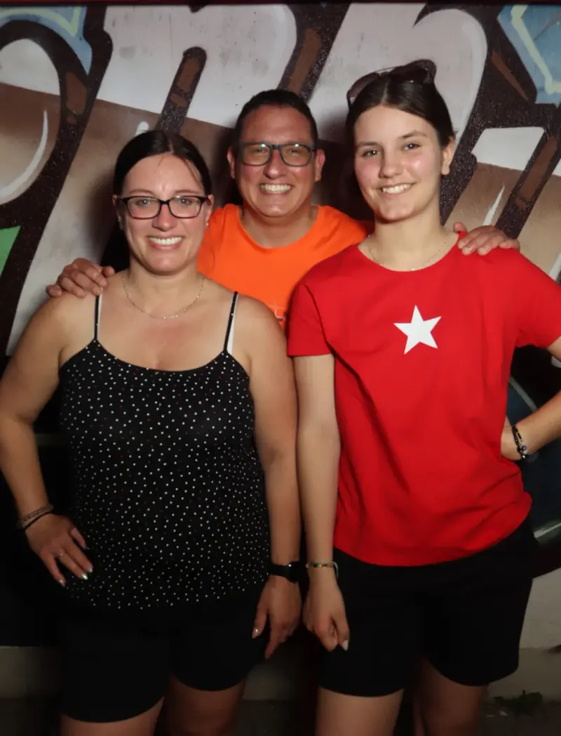 Drei Personen stehen nebeneinander, alle tragen Brillen oder Sonnenbrillen. Im Hintergrund eine Wand mit großem Graffiti.