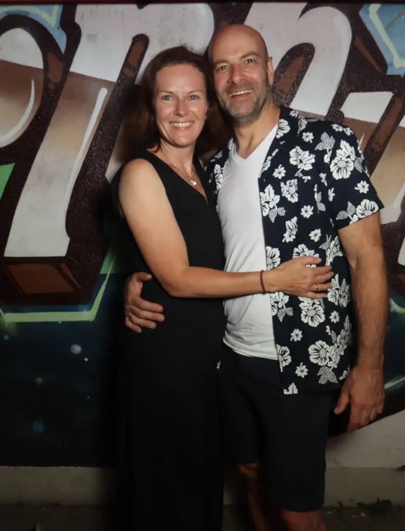 Ein Paar steht eng beieinander, beide lächeln in die Kamera. Im Hintergrund eine Wand mit Graffiti.