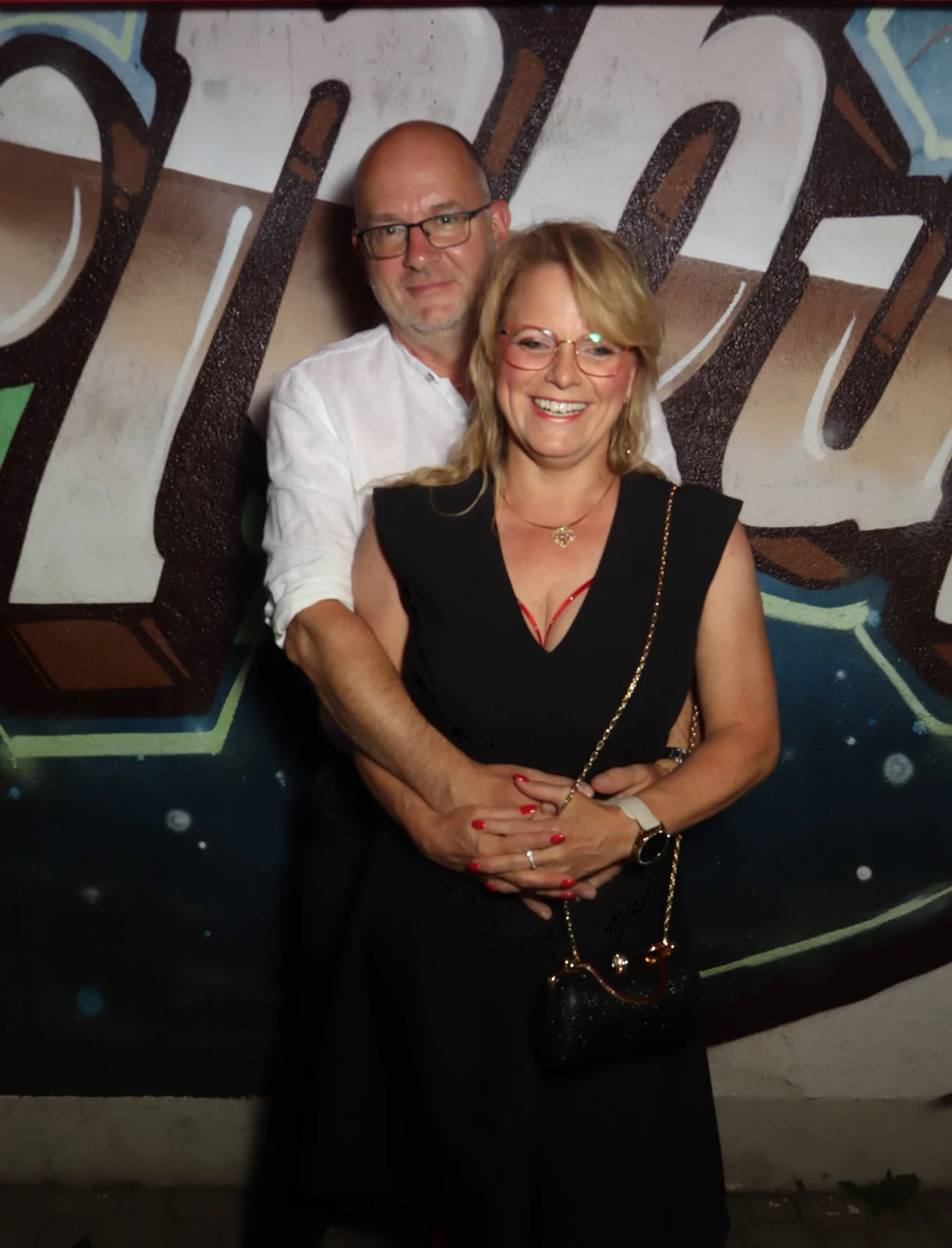 Ein Paar steht umarmt, die Frau trägt ein schwarzes Kleid. Im Hintergrund eine Wand mit Graffiti.