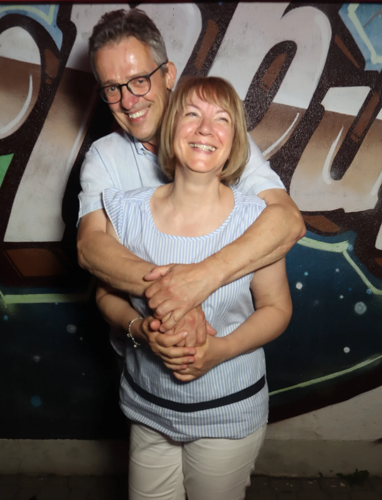 Ein Paar steht umarmt, beide lächeln und tragen helle Kleidung. Im Hintergrund ist eine Wand mit großflächigem Graffiti.