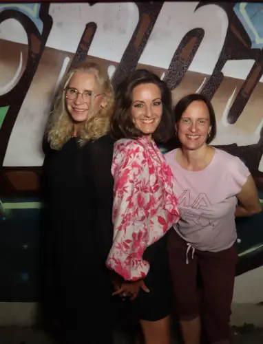 Drei Frauen stehen nebeneinander und lächeln, eine trägt ein florales Oberteil. Im Hintergrund eine Wand mit Graffiti.