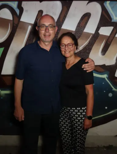 Ein Paar steht nebeneinander, beide tragen Brillen und dunkle Kleidung. Im Hintergrund ist eine bemalte Wand mit Graffiti.
