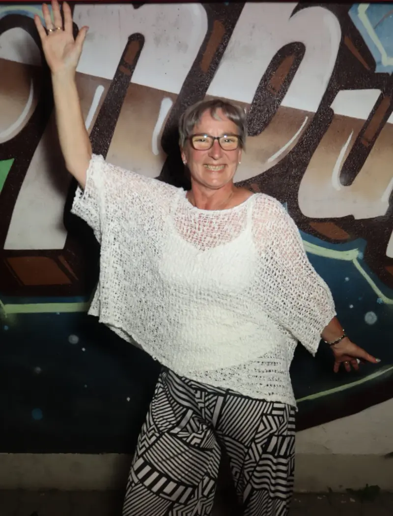 Eine Frau steht mit erhobenen Armen und lächelt, sie trägt ein weißes Oberteil und gemusterte Hose. Im Hintergrund Graffiti.