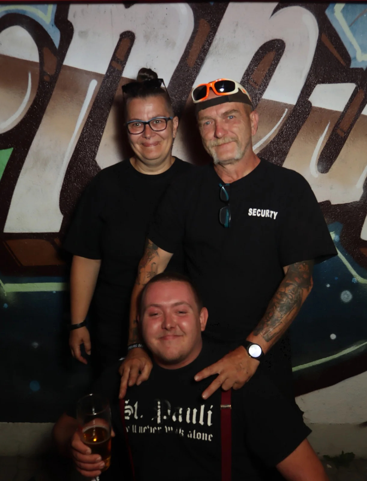 Zwei Personen stehen hinter einer sitzenden Person mit Bierglas, alle tragen schwarze Oberteile. Im Hintergrund Graffiti.