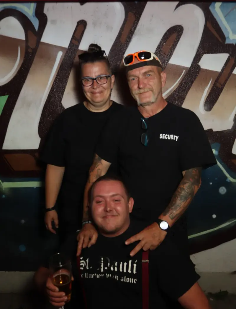 Zwei Personen stehen hinter einer sitzenden Person mit Bierglas, alle tragen schwarze Oberteile. Im Hintergrund Graffiti.