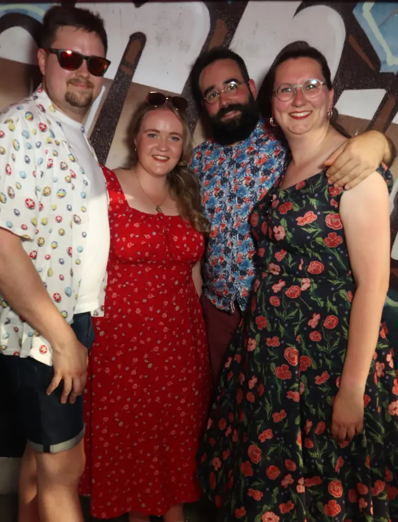 Vier Personen in Sommerkleidung stehen eng zusammen und lächeln. Der Hintergrund ist mit Graffiti gestaltet.