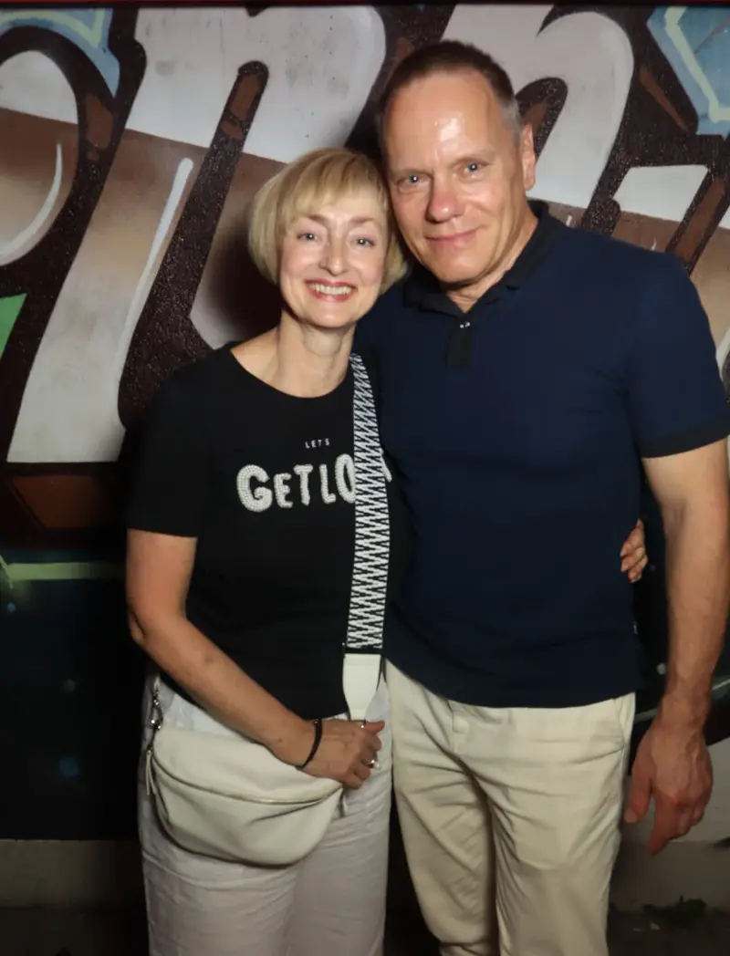 Ein Paar steht eng beieinander, beide tragen dunkle Oberteile und helle Hosen. Im Hintergrund eine bemalte Wand mit Graffiti.