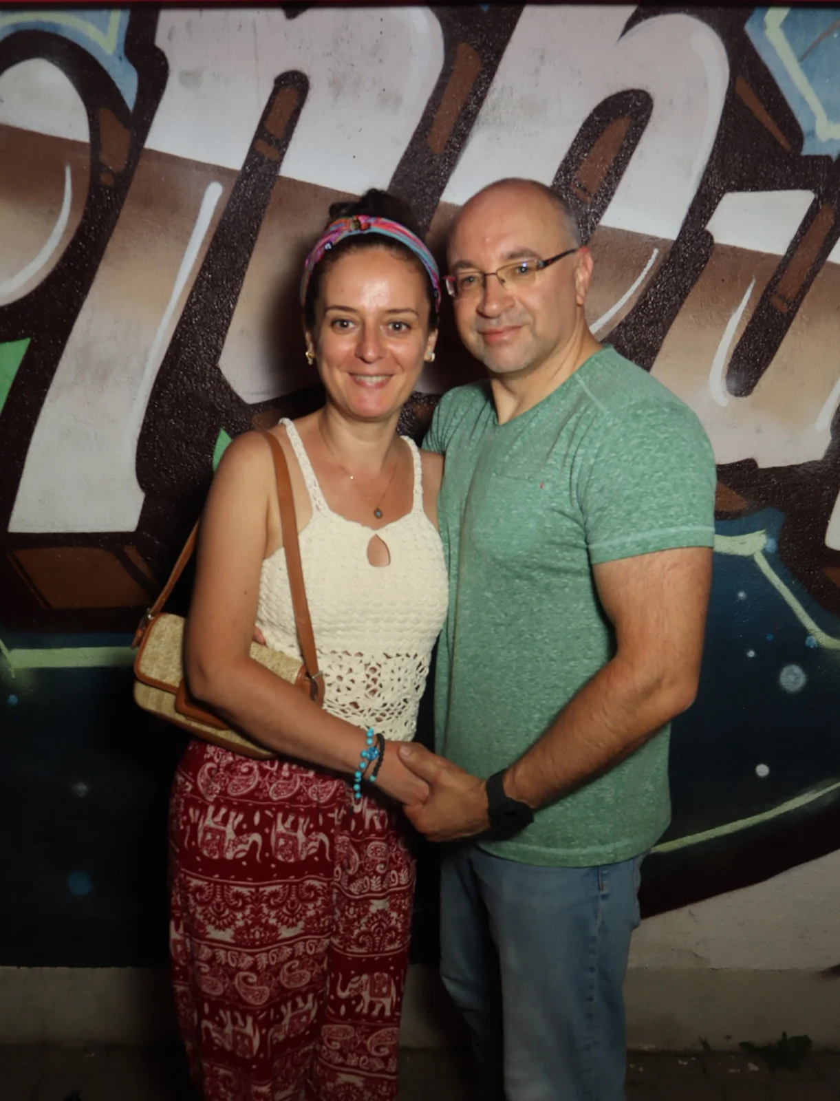 Ein Paar steht nebeneinander und hält sich an den Händen, beide tragen sommerliche Kleidung. Im Hintergrund Graffiti.