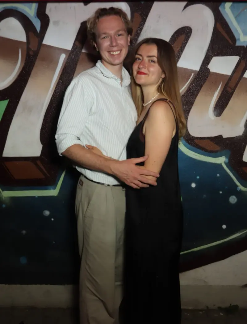 Ein junges Paar steht eng umschlungen, die Frau trägt ein schwarzes Kleid und Perlenkette. Im Hintergrund eine Graffiti-Wand.